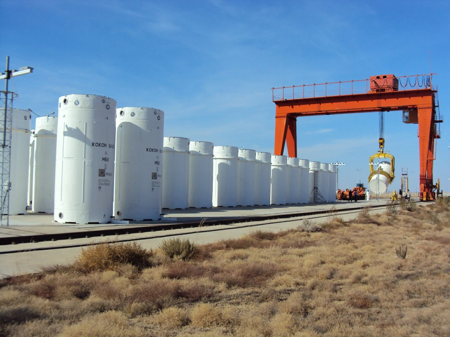 A row of dry-storage casks on a concrete pad, with a crane placing another cask at the far end.